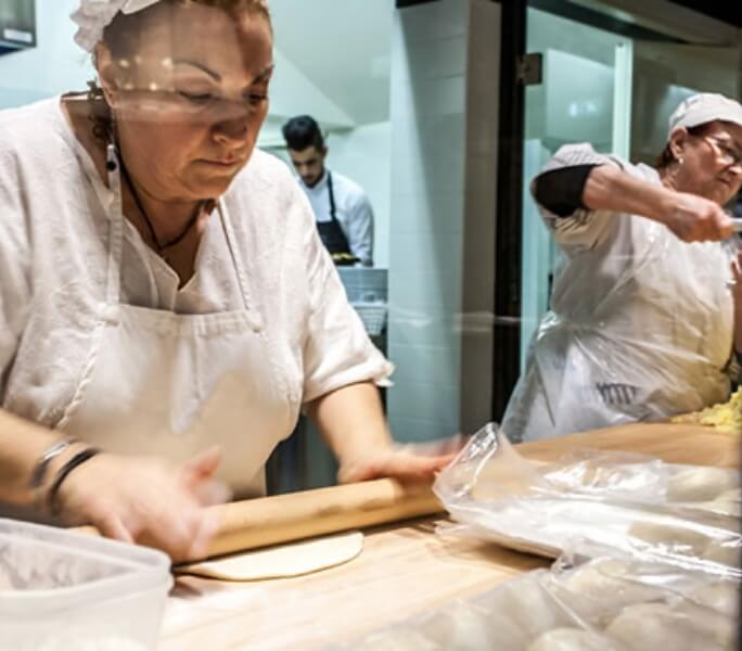 Una donna vestita di bianco con grembiule e cuffia lavora l'impasto della piadina con il matterello su un tavolo da lavoro in legno. Dietro si vedono altri cuochi al lavoro.