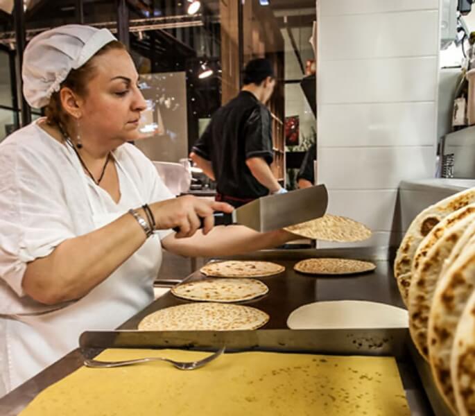 Una donna vestita di bianco con grembiule e cuffia gira con una paletta metallica una piadina che si sta cucinando su di una piastra professionale. Altre piadine si stanno cuocendo sulla piastra.