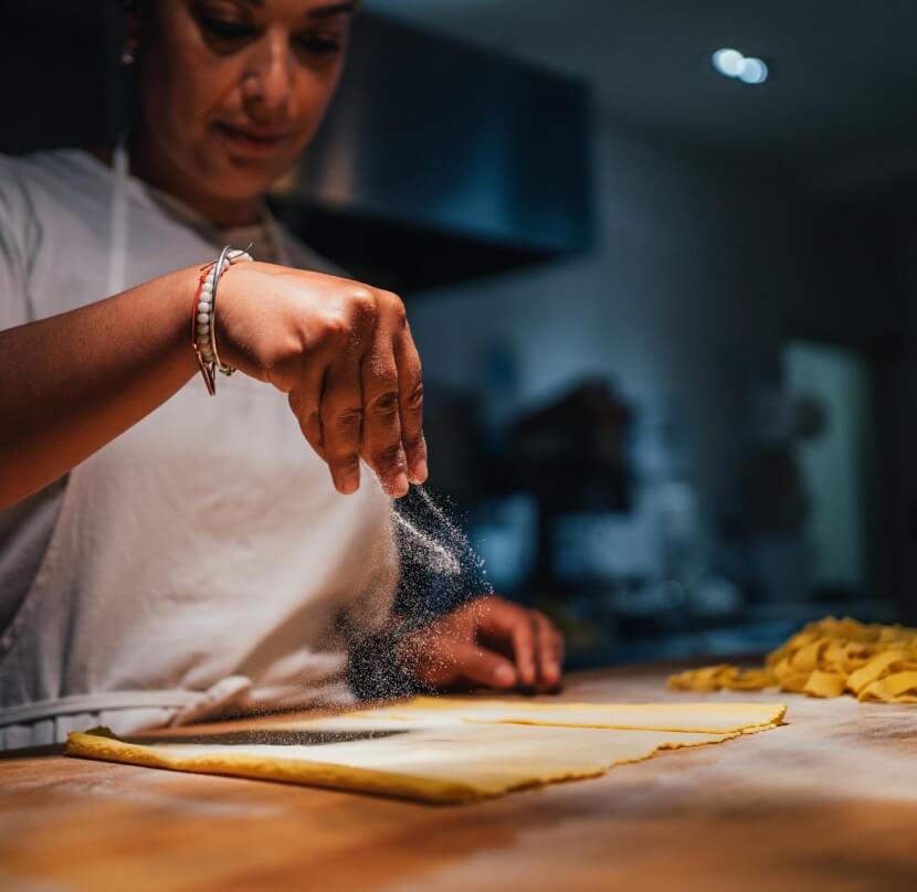 Primo piano di una mano femminile che spande farina sopra una sfoglia di pasta fresca. Nello sfondo si intravedono delle tagliatelle già tagliate